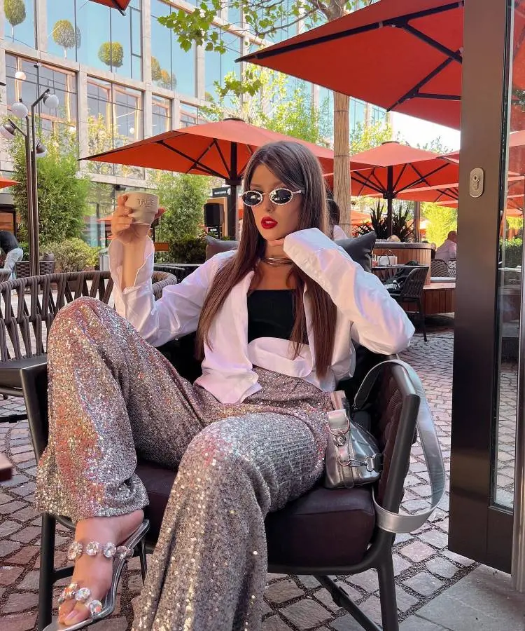 A mulher elegante toma café em um ambiente ao ar livre, vestindo calça brilhante de paetês, sandálias decoradas, camisa branca e top preto. Óculos escuros e batom vermelho complementam o look sofisticado, com guarda-sóis laranja ao fundo.