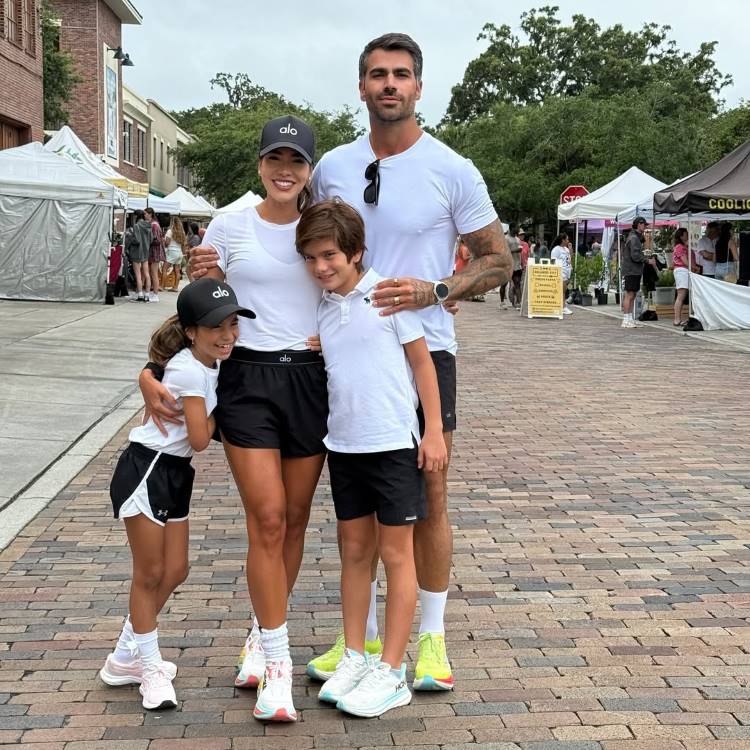 A família de Adriana Sant'Anna e Rodrigão parece feliz em um mercado ao ar livre. Um homem, uma mulher e duas crianças, um menino e uma menina, vestem roupas esportivas brancas e pretas. O homem e a mulher abraçam as crianças.