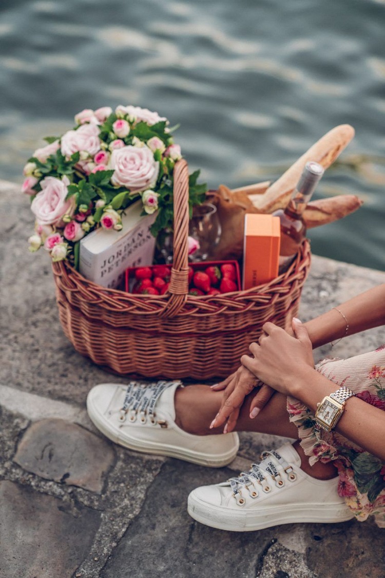 Pernas de pessoa de pele clara usando tênis, ao lado de cesta romântica com vinho, frutas e flores 