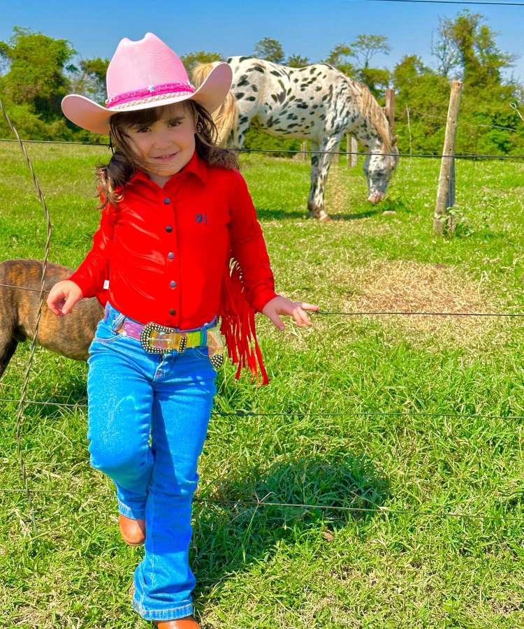 Menina usando roupa country estilo infantil: camisa vermelha com franja, calça jeans, cinto com fivela e chapéu rosa.
