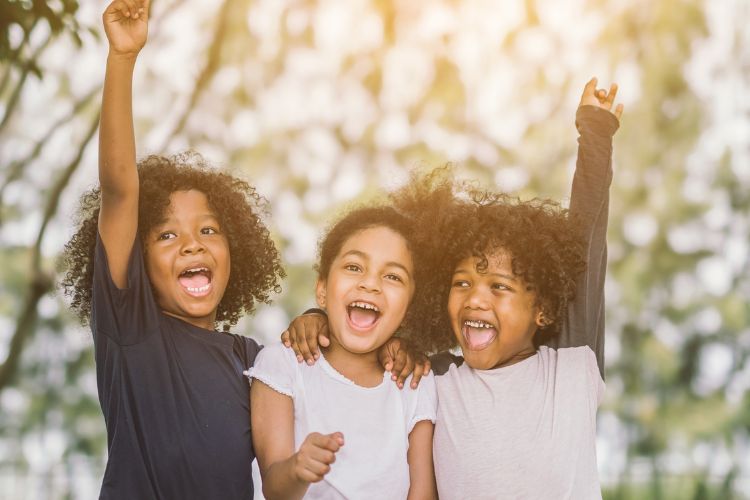 A imagem mostra três crianças sorrindo e demonstrando alegria. Elas estão juntas, com os braços levantados, em um ambiente ao ar livre. A expressão de felicidade delas transmite um momento de diversão e amizade. O fundo desfocado sugere um ambiente natural, possivelmente um parque, com luz suave e agradável.


