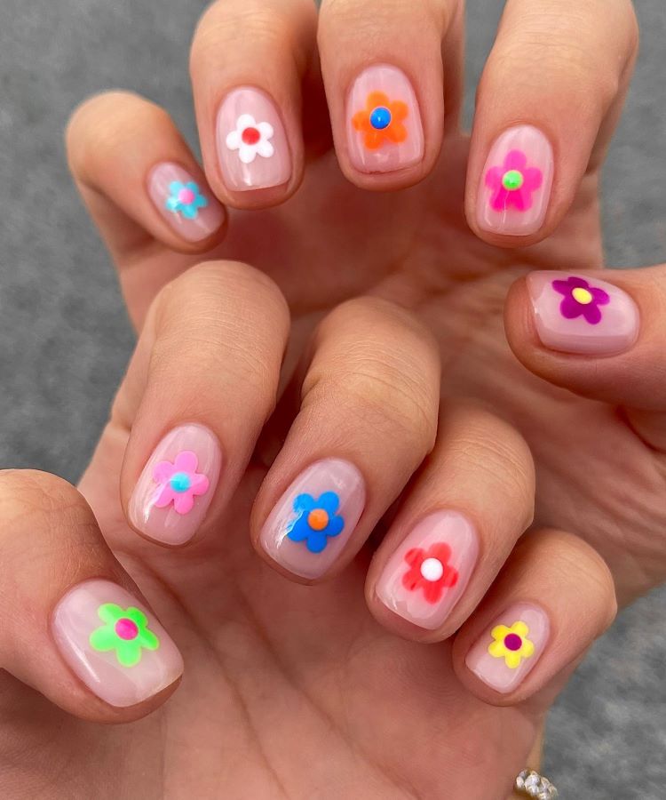 Mãos exibindo estilos de unhas curtas, decoradas com esmalte transparente e pequenas flores coloridas em 3D, incluindo tons de rosa, azul, laranja, verde e roxo. Design alegre e divertido. Fundo cinza desfocado, destacando o contraste vibrante e delicado das unhas.