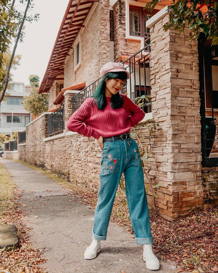 Garota ocidental usando calça jeans bordada, tênis branco, blusa de frio rosa e boina rosa-claro em local com casa decorada com pedras.