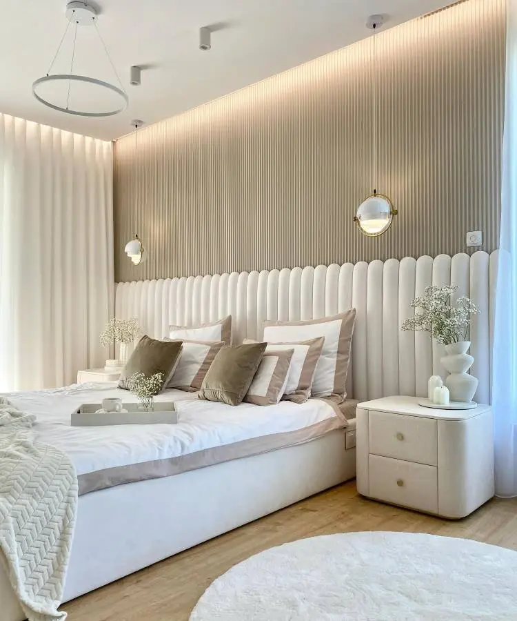 Quarto de casal luxoso, com mesa de canto acolchoada, roupa de cama branco e marrom, cabeceira luxuosa branca estofada
