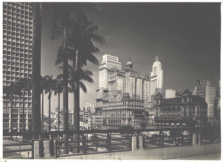 A imagem retrata o centro de São Paulo em meados do século XX, com arranha-céus icônicos, como o Edifício Martinelli e o Altino Arantes, cercados por palmeiras. A composição destaca a modernização e o dinamismo urbano da época.