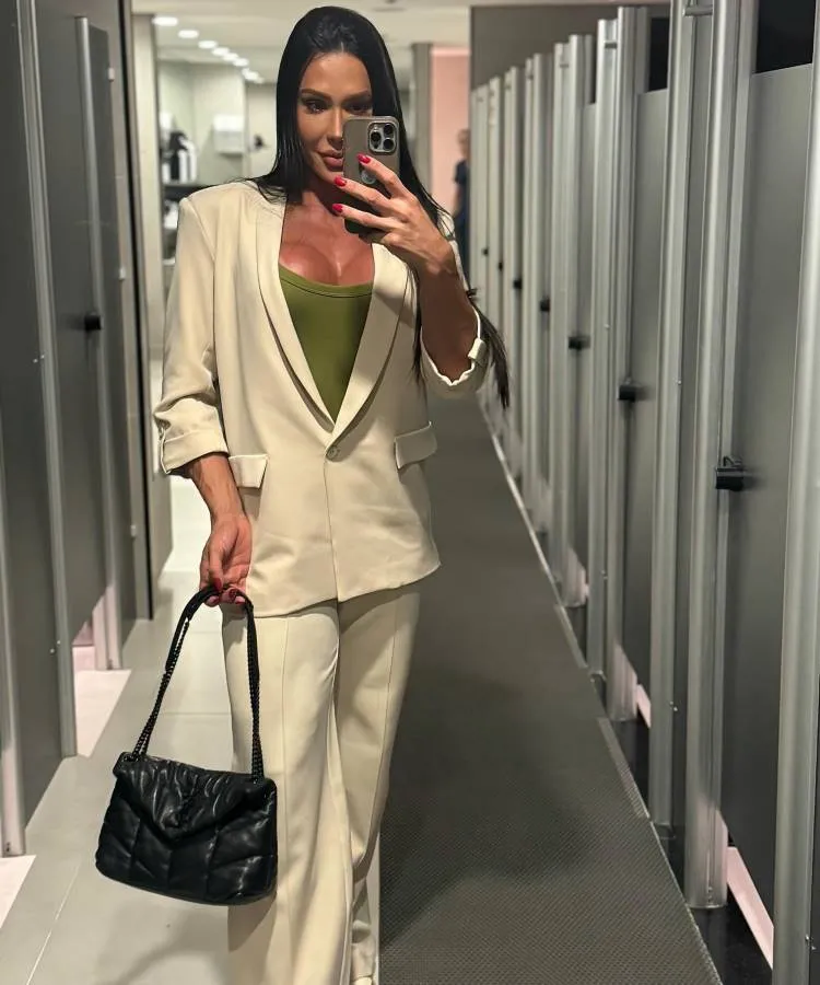 A imagem mostra uma mulher em um provador, tirando uma selfie no espelho. Ela veste um conjunto de blazer e calça em tom claro, com um top verde por baixo, e segura uma bolsa preta. A composição pode ser associada a influenciadores de saúde e estilo que compartilham seu dia a dia.