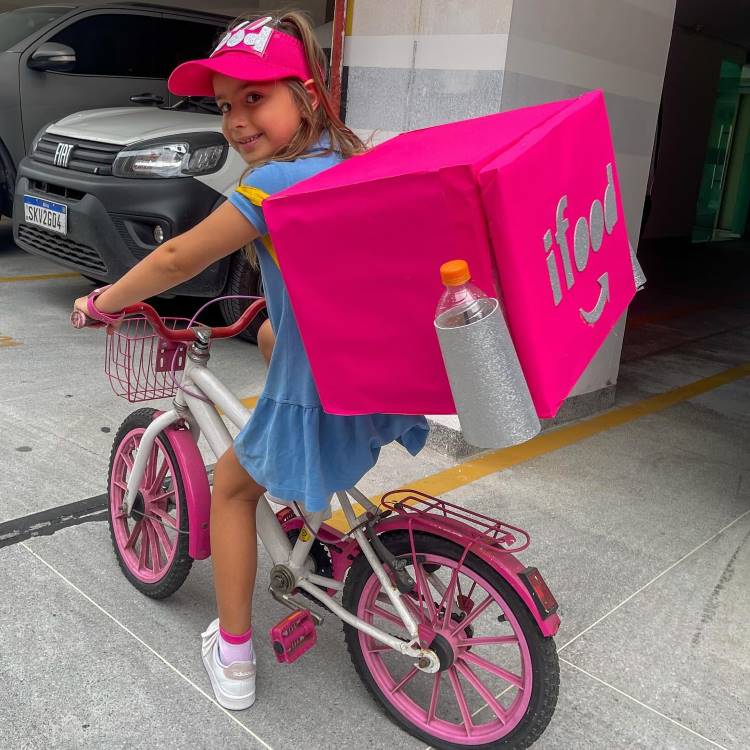A imagem mostra uma menina sorridente em uma bicicleta rosa e branca, usando uma mochila inusitada em forma de caixa rosa brilhante. A mochila se assemelha a uma caixa de entrega de comida, com o logotipo "ifood" visível. A criança veste um vestido azul, boné rosa e tênis brancos. Ela está em uma área que parece ser um estacionamento, com um carro cinza ao fundo.


