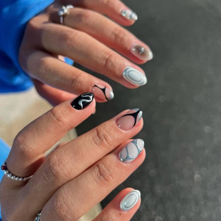 A imagem mostra unhas curtas decoradas com designs abstratos em preto, branco e cinza, incluindo formas onduladas e padrões marmorizados. Os dedos exibem anéis delicados, e o fundo desfocado com luz natural realça o estilo moderno e artístico da nail art.