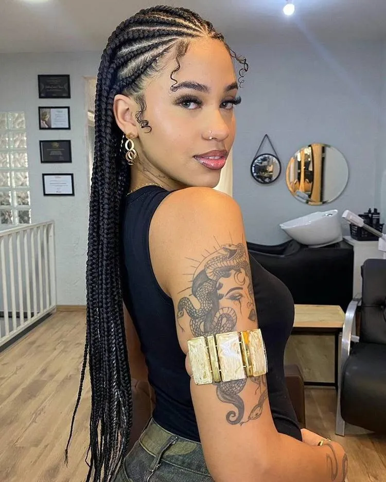 Mulher com trança nagô longa, até o bumbum. Ela está usando regata preta e calça jeans em salão de beleza