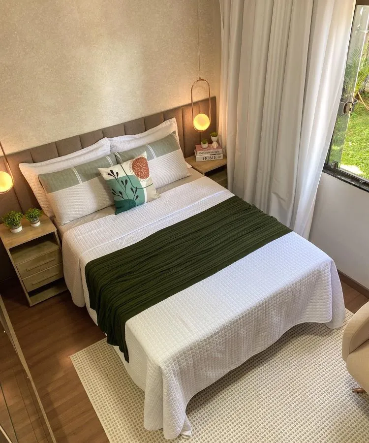 A imagem mostra uma decoração fácil para quarto com uma cama de casal, lençóis brancos e uma manta verde. Há almofadas decorativas, duas mesinhas de cabeceira com abajures pendentes e um tapete claro. A combinação de cores neutras e iluminação natural cria um ambiente acolhedor e relaxante.

