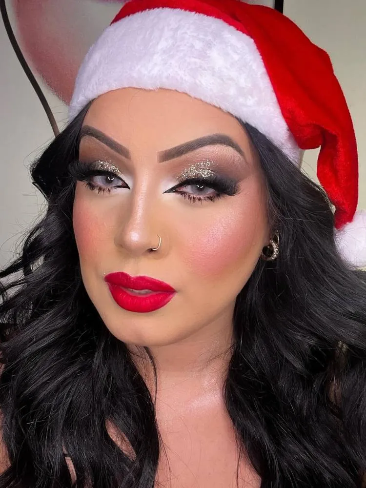 Mulher de pele clara com gorro natalino na cabeça usando maquiagem carregada nos olhos, com brilho prata, cílios postiço e delineado, além de batom vermelho. 