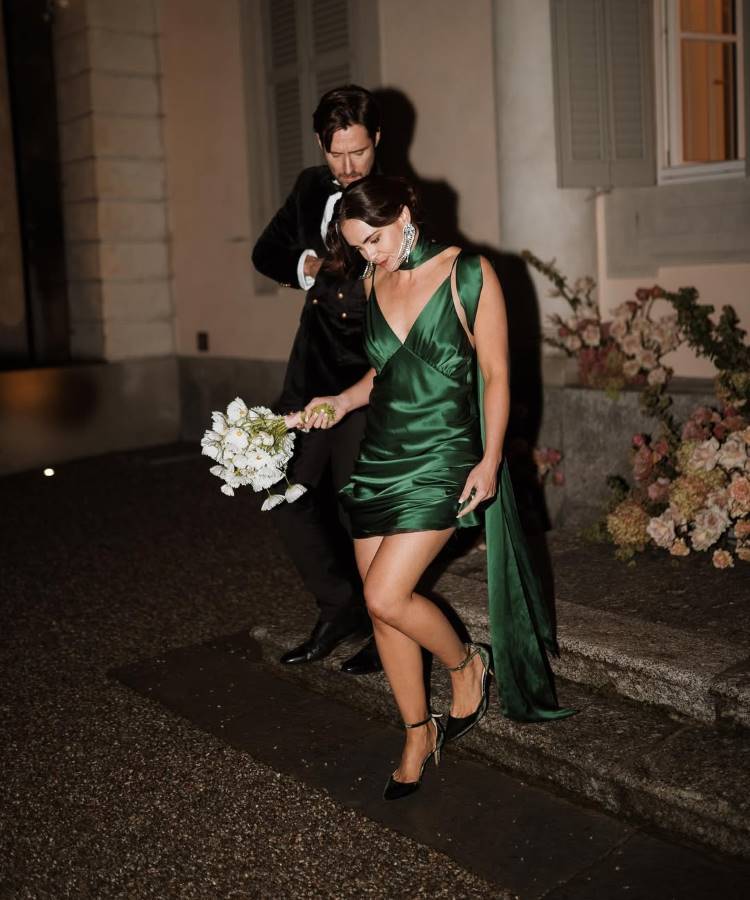 Uma noiva com um vestido verde-esmeralda curto de seda, gola lenço e cauda longa, segura um buquê branco e olha para o noivo de terno escuro. Eles caminham em uma área externa noturna, com flores ao fundo.







