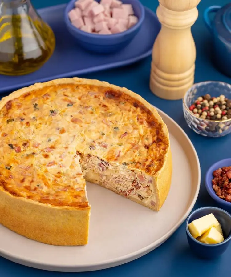 Uma quiche parcialmente cortada exibe seu recheio cremoso recheado de presunto e ervas sobre um prato bege. Ao redor, ingredientes como cubos de presunto, manteiga, pimenta em grãos e azeite, sobre uma mesa azul, completam a cena culinária.