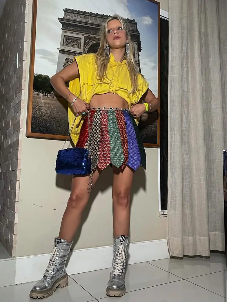 Mulher de pele clara de cropped amarelo e saia de gravatas coloridas, além de coturno prata 