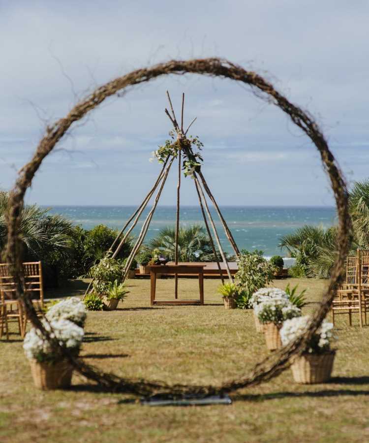 Foto de cerimônia ao ar livre com arco de galhos decorado por flores brancas. Ao fundo, altar rústico de madeira em frente ao mar. Cadeiras e cestos com flores compõem o ambiente natural e romântico.