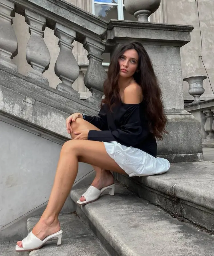 Uma mulher com cabelos castanhos longos e ondulados está sentada em uma escadaria de pedra, vestindo um top preto de ombro a ombro e uma saia branca. Ela usa sandálias brancas de salto.