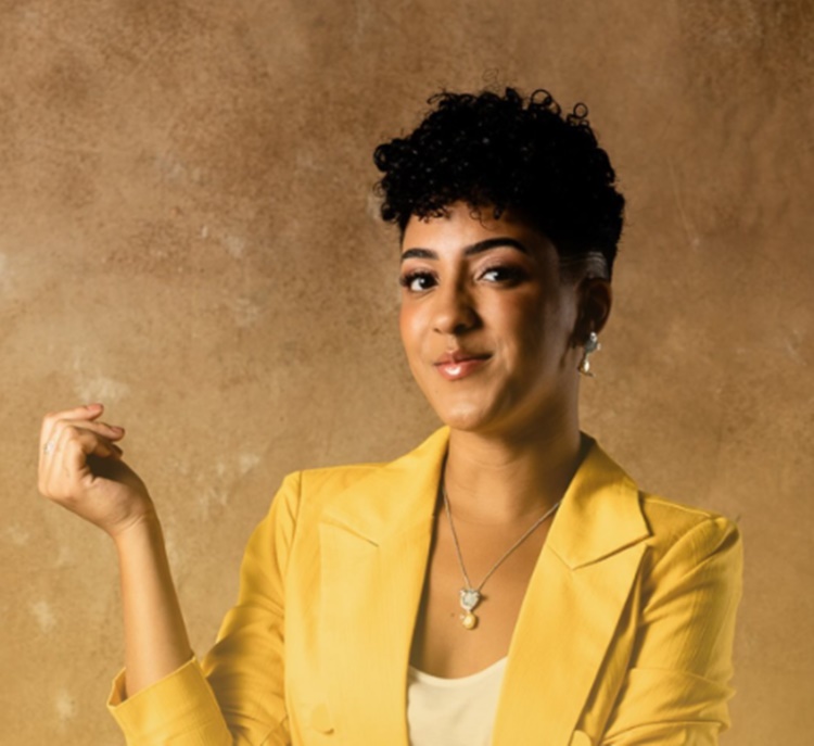 A imagem mostra Nath Finanças sorrindo, com cabelo curto e encaracolado. Ela usa um blazer amarelo e um colar discreto. O fundo é de cor neutra, realçando a expressão confiante e amigável.


