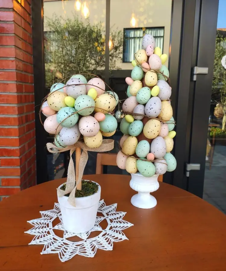 Mesa decorada com arranjos temáticos de Páscoa, contendo árvores feitas de ovos coloridos em tons pastel com detalhes pintados em preto. Uma base branca e outra de vaso rústico com laço decorativo, sobre toalha de crochê branca. Fundo externo.