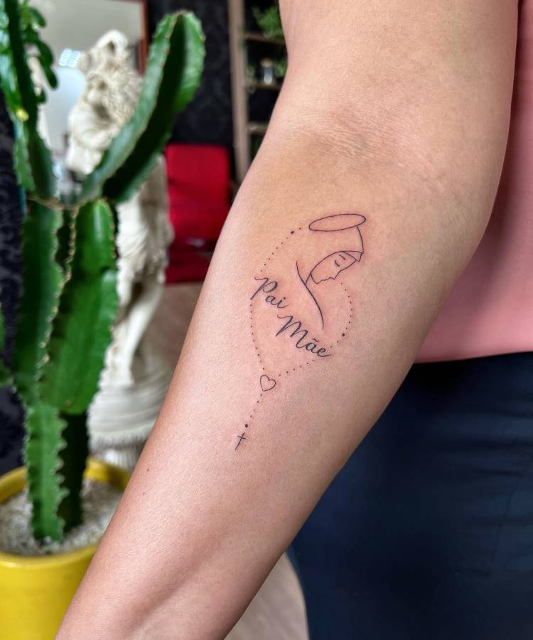 A imagem exibe uma tatuagem no antebraço de uma pessoa, apresentando o contorno de uma figura religiosa com auréola, cercada por uma linha pontilhada em forma de coração. As palavras "Pai Mãe" estão escritas em cursiva. Uma cruz e um pequeno coração finalizam o desenho. Um cacto é visível no fundo.







