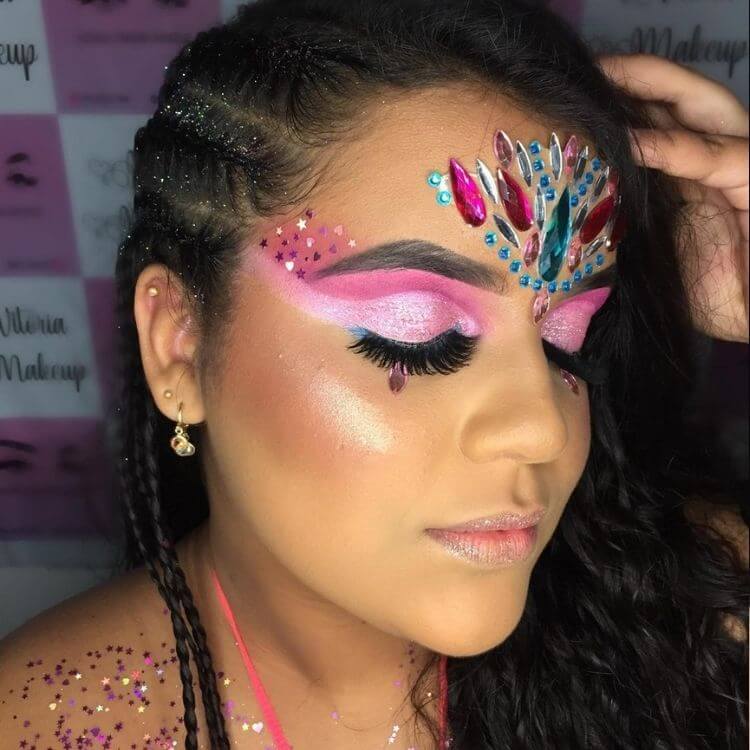 Mulher de pele morena com maquiagem rosa até o cabelo, glitter em estrela e coração, além de pedrarias azuis e rosas na testa 