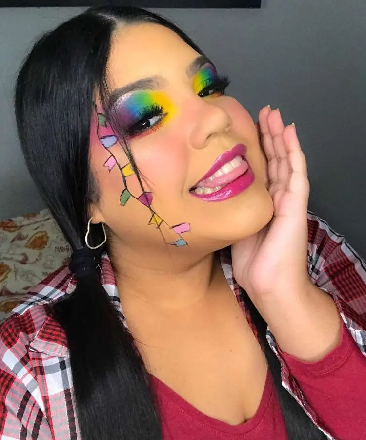 Uma mulher com maquiagem colorida para Festa Junina, exibindo uma sombra de olhos em tons de arco-íris e um desenho de bandeirinhas coloridas descendo pela lateral do rosto. Seus lábios são de um rosa vibrante.
