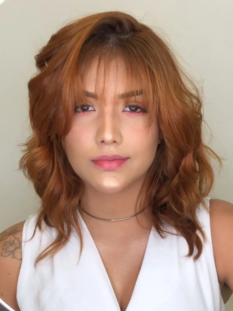 Mulher ruiva de pele clara com rosto arredondado, usando blusa branca e franja coreana no cabelo médio