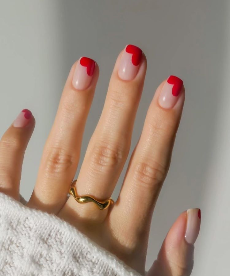 A imagem mostra uma mão com unhas decoradas em um estilo moderno. As unhas têm uma base em tom nude e são adornadas com um detalhe vermelho em formato de coração na ponta. A mão está usando um anel dourado com um design ondulado. A pessoa veste uma peça de roupa branca, provavelmente um suéter de malha. A iluminação suave destaca os detalhes das unhas e do anel.