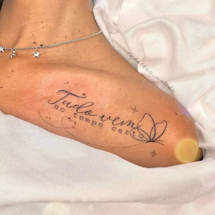 Mulher de pele clara com corrente prateada e tatuagem de frase escrito "tudo vem no tempo certo" com borboleta.