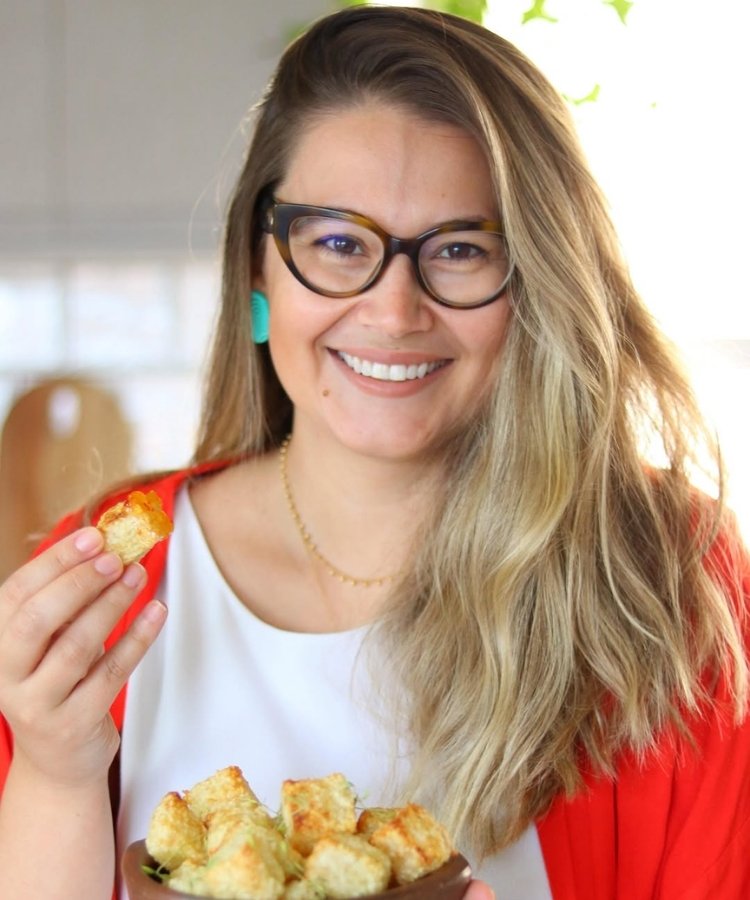 Foto de Marilia Archangelo usando camiseta branca, casaco laranja e segurando dadinhos de tapioca
