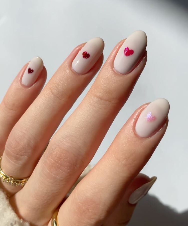A imagem mostra uma mão com unhas esmaltadas em um tom claro, cada uma decorada com um pequeno coração de cor vermelha ou rosa. As unhas têm um formato oval e estão bem cuidadas. A mão está usando anéis dourados finos. A iluminação é natural e destaca o brilho suave do esmalte e dos detalhes dos corações.