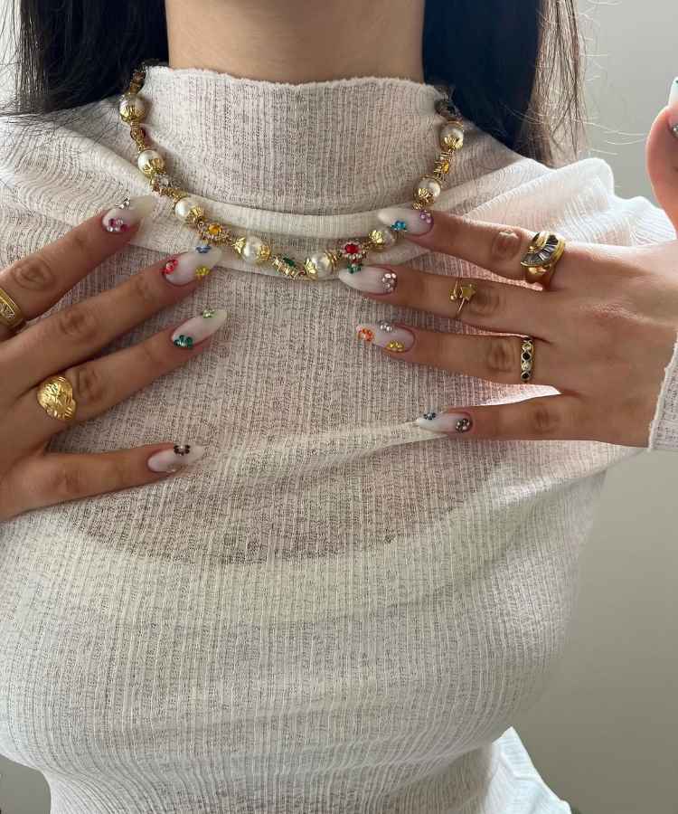Mulher vestindo blusa branca texturizada, exibe unhas longas decoradas com cores vibrantes e pedrarias. Usa colar dourado com pérolas e anéis variados. As mãos estão posicionadas sobre o peito, realçando os acessórios e o design das unhas contra o tecido claro.