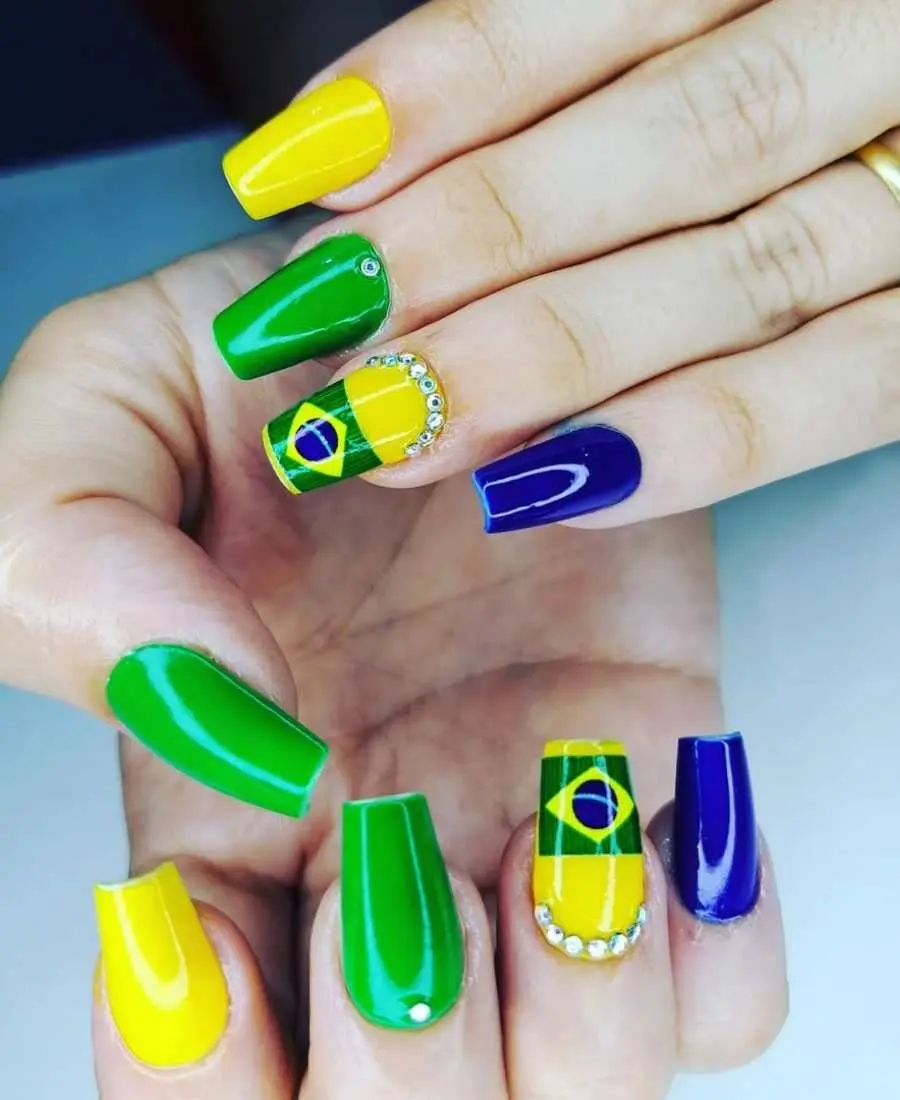 Imagem de mão decoradas com esmalte.  Cores usadas, verde, amarelo, azul e branco. Polegar: verde. Indicador: amarelo. Dedo Médio, verde com 1 pedrinha. Anelar: amarelo com bandeira + várias pedrinhas. Mínimo: azul.