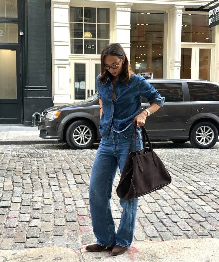 A imagem mostra uma mulher vestindo um conjunto jeans tendência 2025, com camisa de lavagem média e calça wide leg. O look casual é complementado por sapatos marrons, uma bolsa grande de couro e óculos, criando um estilo moderno e prático.
