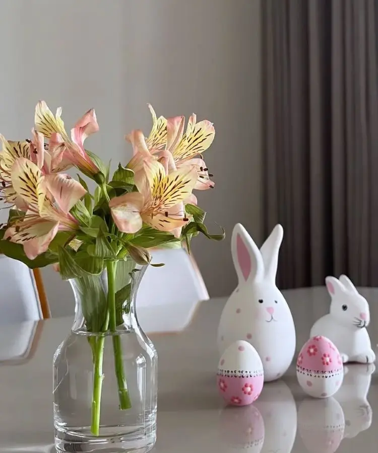 Mesa decorada com enfeites de Páscoa, incluindo coelhos brancos e ovos decorativos rosa com detalhes delicados. Ao lado, um vaso de vidro com flores amarelas e rosadas. Ambiente moderno e iluminado, com cortina cinza ao fundo.