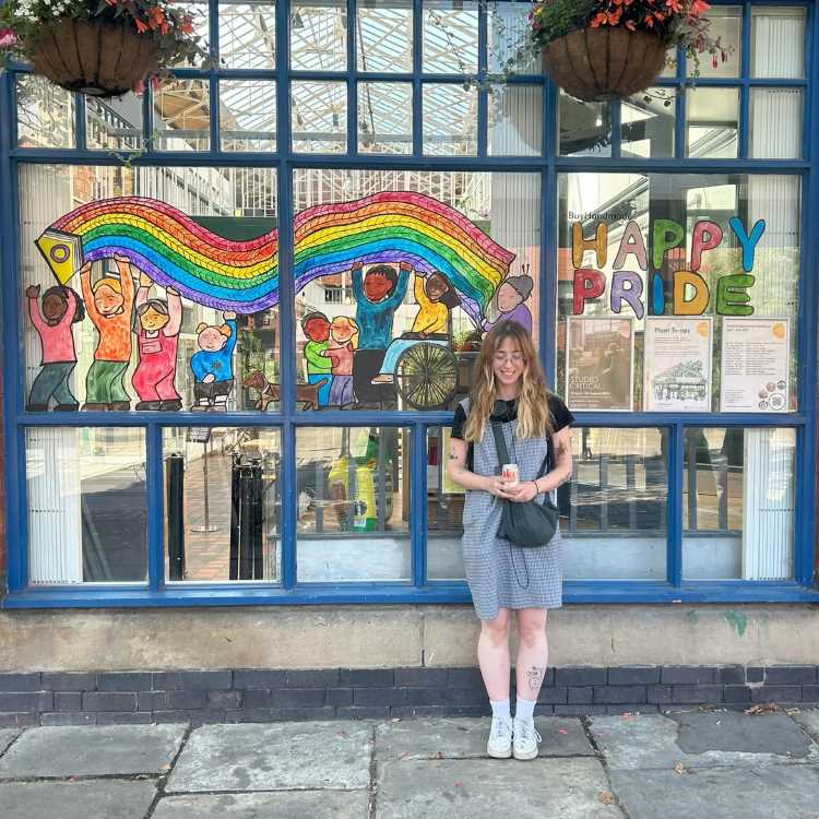 Fachada de vidros com ilustração de arco-íris, crianças comemorando e a frase "Happy Pride" em letras coloridas. Mulher sorri em frente à vitrine, vestindo roupas casuais. Vasos pendurados e cenário urbano criam ambiente alegre e inclusivo.