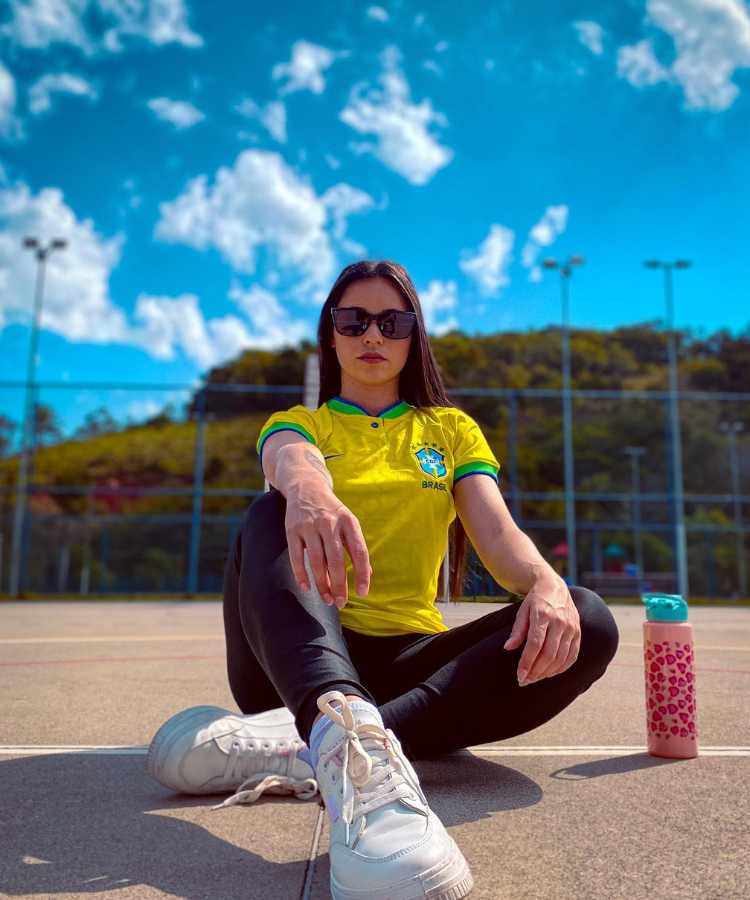 Imagem com fundo de quadra. No centro, mulher usando camisa do Braisl, legging preta + tênis branco + óculos de sol