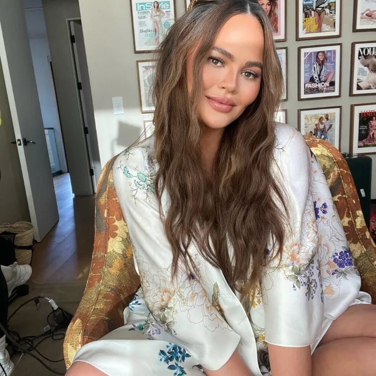 Chrissy Teigen posa em uma poltrona, ela tem cabelos longos castanhos em camadas e usa um kimono branco de flores