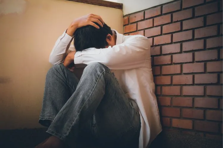 Homem sentado no chão, encostado em uma parede de tijolos e outra bege, com as mãos na cabeça em sinal de desespero ou tristeza profunda. A postura e o ambiente sugerem angústia, talvez relacionada a problemas pessoais ou impactos da ludopatia.