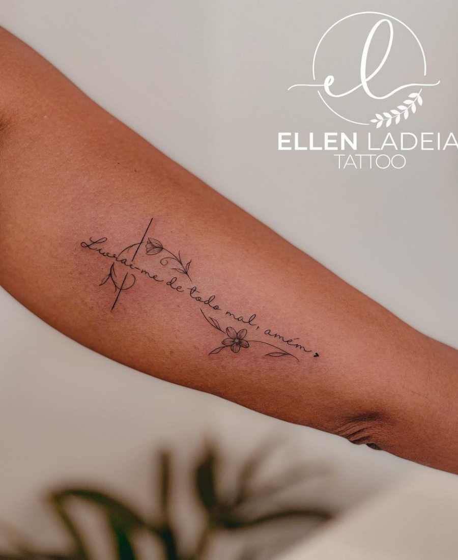foto de tatuagem religiosa com frase e flores no braço de uma pessoa.