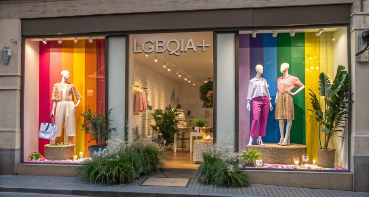 Fachada de loja com vitrine temática LGBTQIA+, decorada com cores de arco-íris em painéis de fundo. Manequins exibem roupas elegantes. Plantas em vasos e iluminação suave completam o ambiente. O interior luminoso e acolhedor é visível ao fundo.
