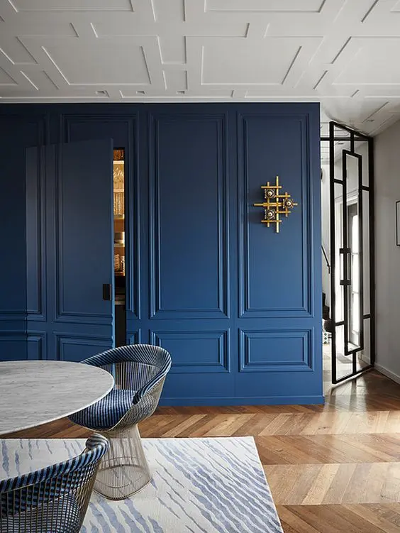 Sala de jantar com piso de madeira, mesa oval, cadeiras azuis, tapete bege e azul e parede azul com moldura de mesma cor 