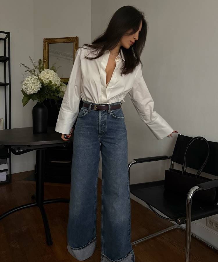 Mulher estilosa posa em ambiente minimalista, usando camisa branca de tecido leve com jeans de cintura alta e barra dobrada. Cabelo solto, acessórios discretos e ambiente sofisticado ao fundo. Ótima inspiração para looks de trabalho casuais e elegantes.





