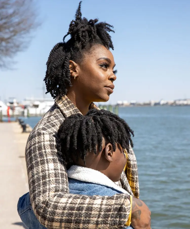 Mãe e filho, de etnia negra, observam a água em um dia ensolarado. A mulher olha para o horizonte, enquanto o menino se apoia nela. A imagem transmite tranquilidade e união familiar.