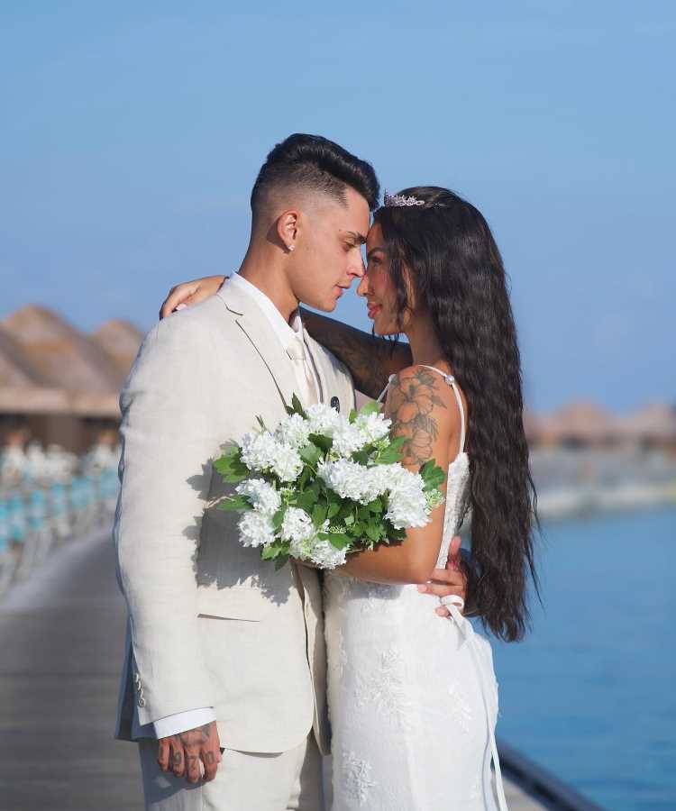 Casal Pamella Rodrigues e Victor Lunardi em momento romântico à beira-mar. Noiva usa vestido branco rendado, com tiara e segura buquê de flores brancas. Noivo veste terno claro. Cenário paradisíaco, com palafitas e céu azul ao fundo, transmite elegância e serenidade.

