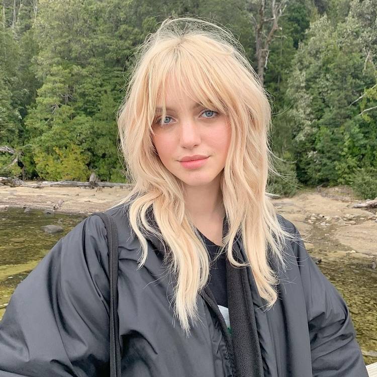 Mulher com cabelo loiro claro e franja, usando jaqueta preta acolchoada. Ela está ao ar livre, em um cenário natural com árvores e um rio ao fundo. A expressão é suave e confiante, destacando-se na paisagem verde e serena.