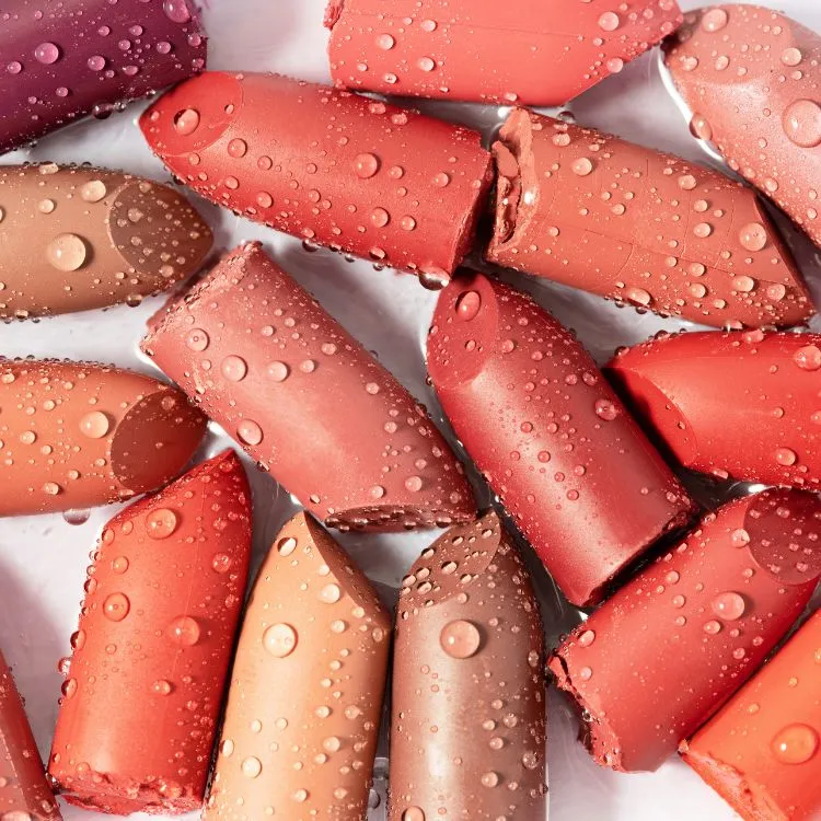 A imagem mostra várias pontas de batons dispostas juntas, com diferentes tons que variam do rosa claro ao vermelho profundo e roxo. A superfície dos batons parece lisa, com gotas de água espalhadas, sugerindo um ambiente fresco ou úmido.

