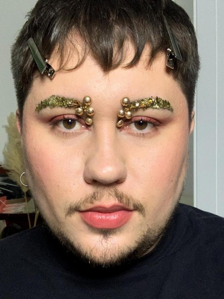 Homem de pele clara com glitter dourado na sobrancelha e pérolas também douradas