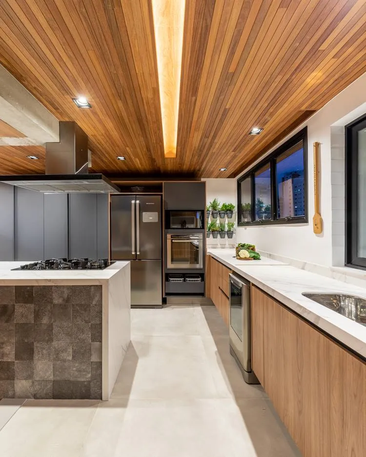 Vista da Cozinha de apê de 187 m² decorada pelo arquiteto Pietro Terlizzi com direção à geladeira de 2 portas em cor inox, e móvel com forno convencional e microondas. Na parede da bancada, 6 vasos pequenos de plantas