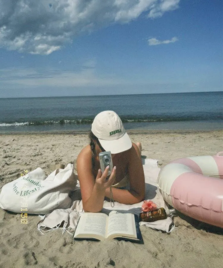 Uma mulher deitada na areia da praia, lendo um livro. Ela usa um boné branco e segura uma câmera. Ao seu lado, há uma bolsa de praia e um protetor solar. O mar e o céu azul com nuvens completam a cena.




