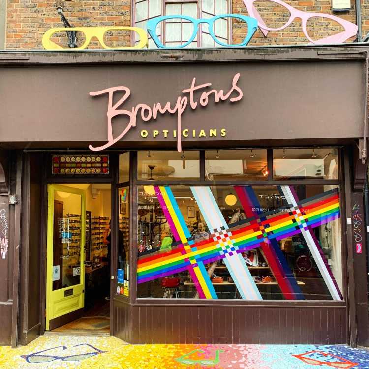 Fachada da ótica Bromptons, com logotipo em rosa e amarelo. A vitrine exibe faixas coloridas em tema arco-íris, celebrando diversidade. Piso decorado com óculos pintados em cores vibrantes. Interior iluminado destaca armações expostas em prateleiras. Atmosfera acolhedora e moderna.